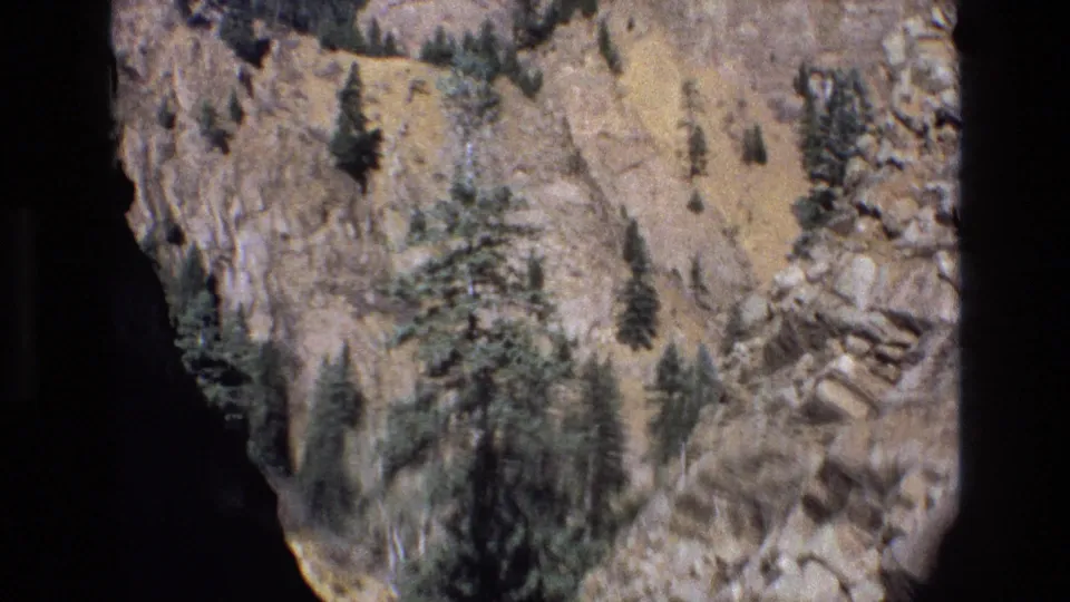 1973 Colorado 8mm frame showing a rocky mountain slope with scattered evergreen trees, dry scrub, blue sky, and dark gate edges from the home-movie scan.