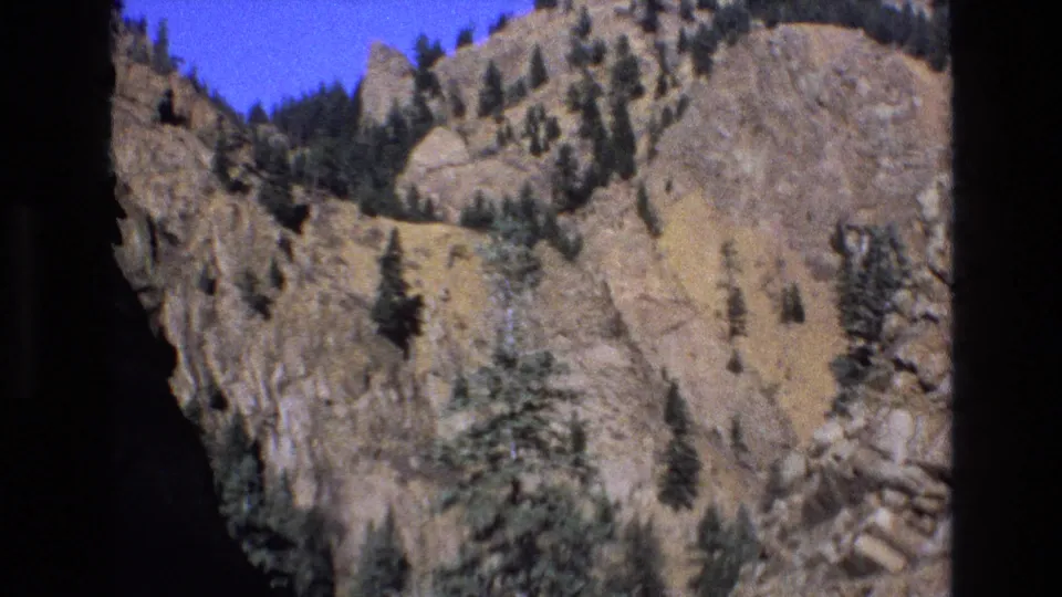 1973 Colorado home-movie frame with a sunlit rock face, sparse evergreens, warm dry hillside tones, saturated blue sky, and pronounced dark vignetting.