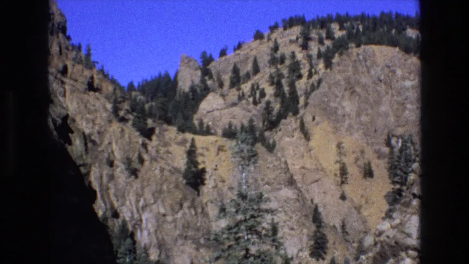 1973 Colorado 8mm frame widening to show rugged cliffs, scattered evergreens, dry mountain slopes, blue sky, and handheld home-movie framing.