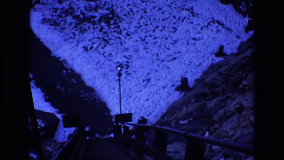Blue-tinted 1973 Colorado 8mm frame showing a shadowed mountain road or overlook structure between rocky slopes, with pale ground, rail-like edges, and dark film borders.