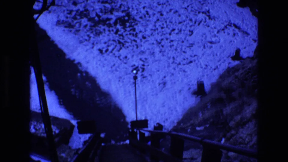 Blue-shifted 1973 Colorado 8mm frame showing snow-covered ground, a dark bridge or walkway edge, and a small lamp post in a nighttime mountain setting.