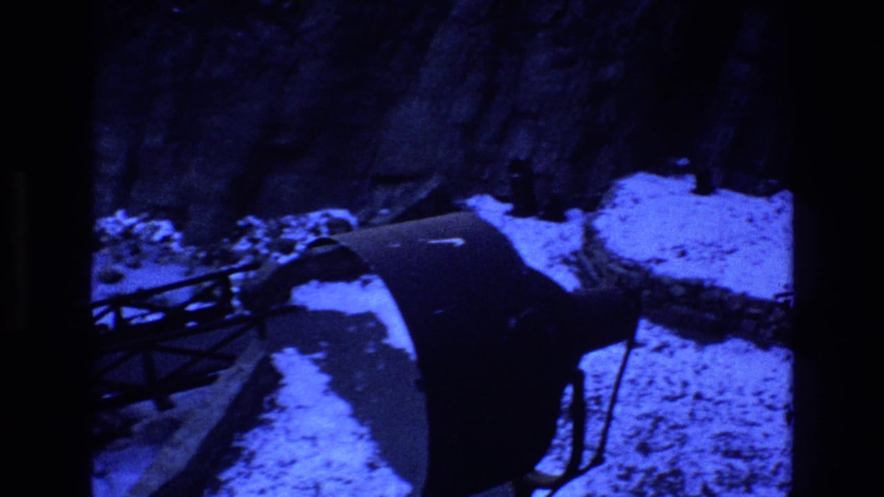 1973 Colorado 8mm frame looking down at a snow-lined riverbank beside a rocky canyon wall, with part of a dark metal bridge crossing the lower left of the view.