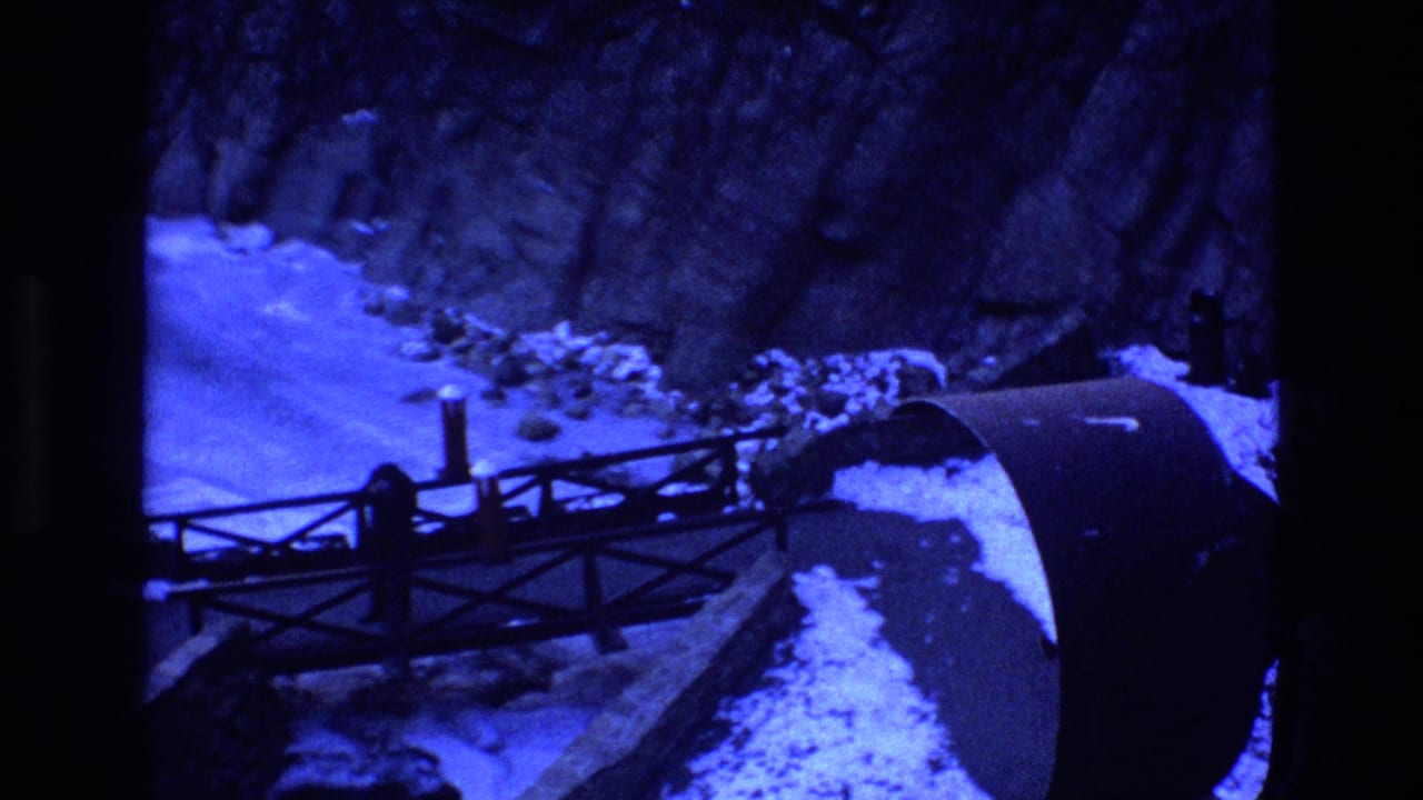 Metal bridge spans a blue-tinted snowy waterway below steep rock walls.