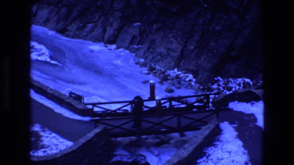 1973 Colorado 8mm frame looking down on a river bend bordered by snow and canyon rock, with a dark bridge railing crossing the foreground.