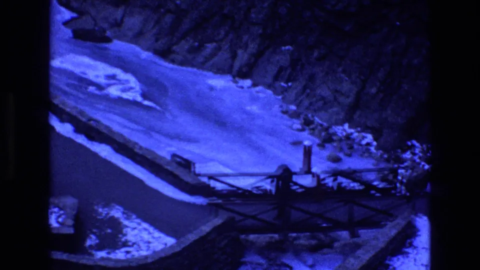 1973 Colorado 8mm frame of a snow-edged river or waterway below steep canyon rock, with a dark metal bridge crossing the lower foreground under a strong blue cast.