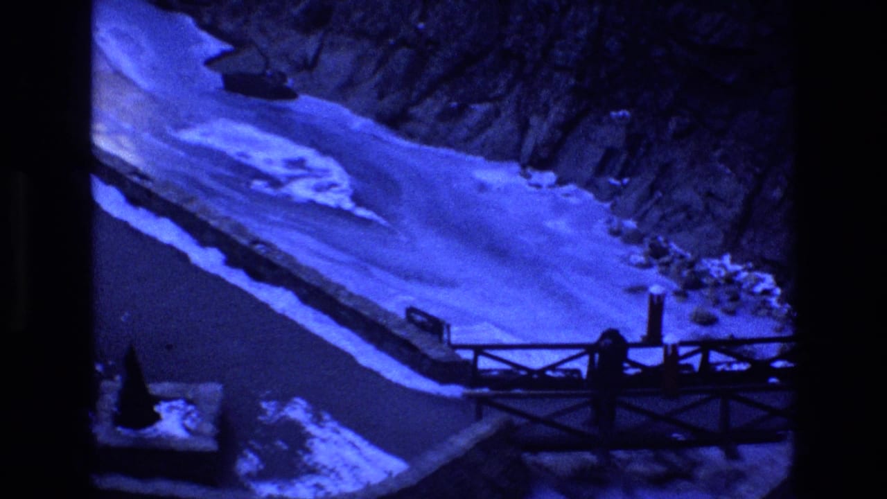 1973 Colorado 8mm frame showing a blue-shifted snow-covered path, rustic wooden railing, steep rocky slope, and dark canyon shadows.