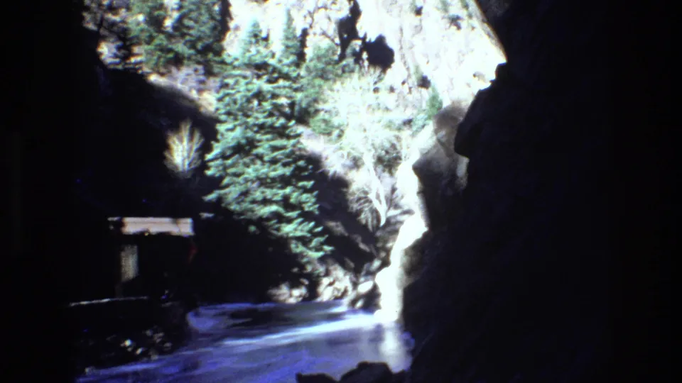 1973 Colorado home-movie frame looking through a shaded rocky gorge toward sunlit evergreens, snow, and a small distant figure on the path.