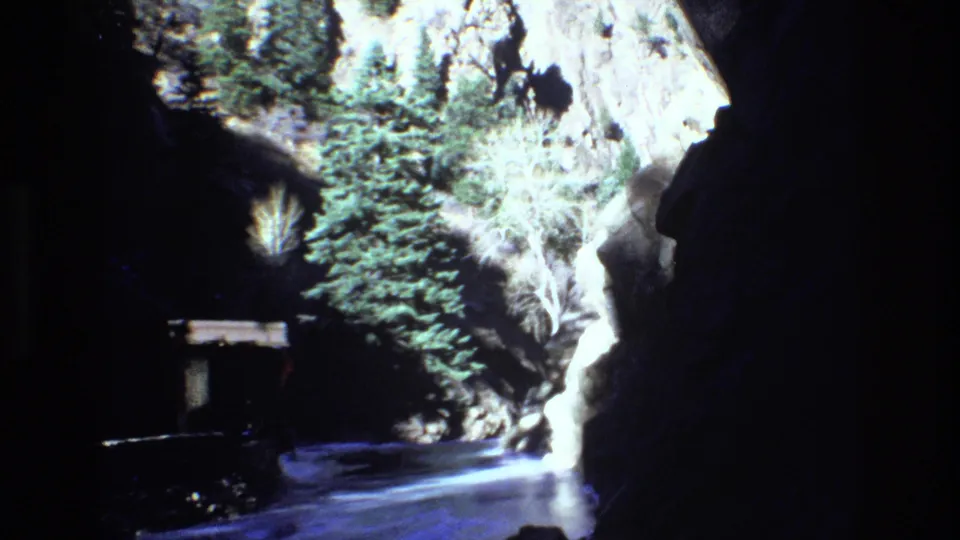 1973 Colorado 8mm frame of a shadowed canyon passage with evergreen trees, pale rock, snow on the ground, and a small human figure near the path.