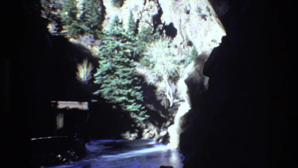 1973 Colorado home-movie frame showing a narrow snow-lined path between dark rock walls, sunlit pale stone, evergreens, and a distant person.
