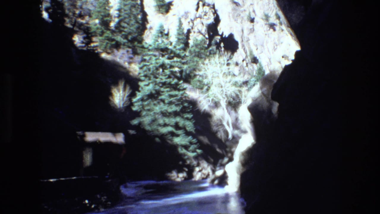 1973 Colorado 8mm frame showing a rocky canyon passage with evergreen trees, bright water, and a small shelter-like structure near the shaded edge.