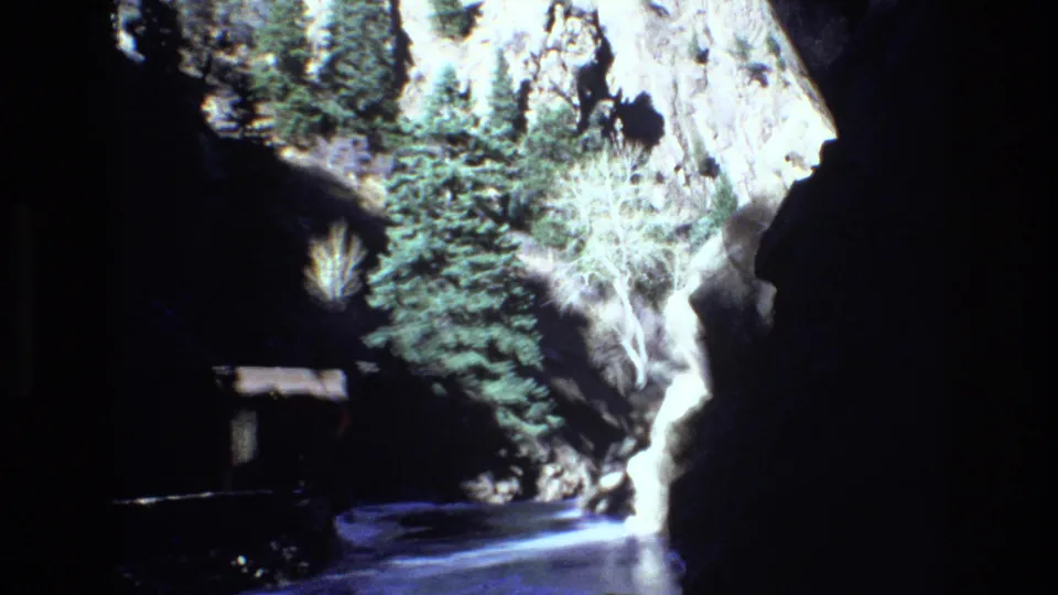 1973 Colorado 8mm frame showing a rocky canyon passage with evergreen trees, bright water, and a small shelter-like structure near the shaded edge.