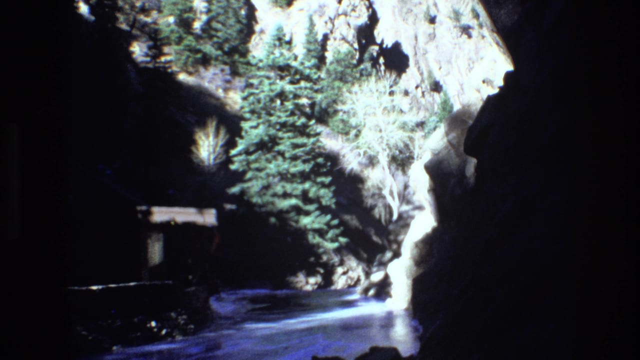 1973 Colorado 8mm frame looking into a narrow rocky canyon with evergreen trees, reflective water, and a small structure partly visible along the left side.