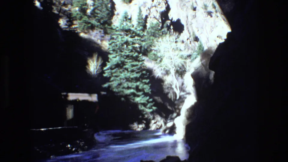 1973 Colorado 8mm frame looking into a narrow rocky canyon with evergreen trees, reflective water, and a small structure partly visible along the left side.