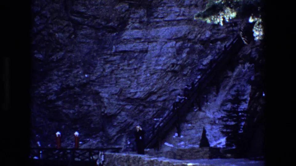1973 Colorado 8mm frame showing a steep rocky hillside with a wooden stairway or walkway against the cliff and distant visitors near railings below.