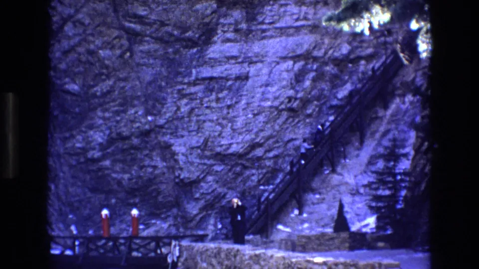 1973 Colorado 8mm frame of a rocky cliff face with a steep wooden stairway, railings, and distant people standing near the base of the slope.