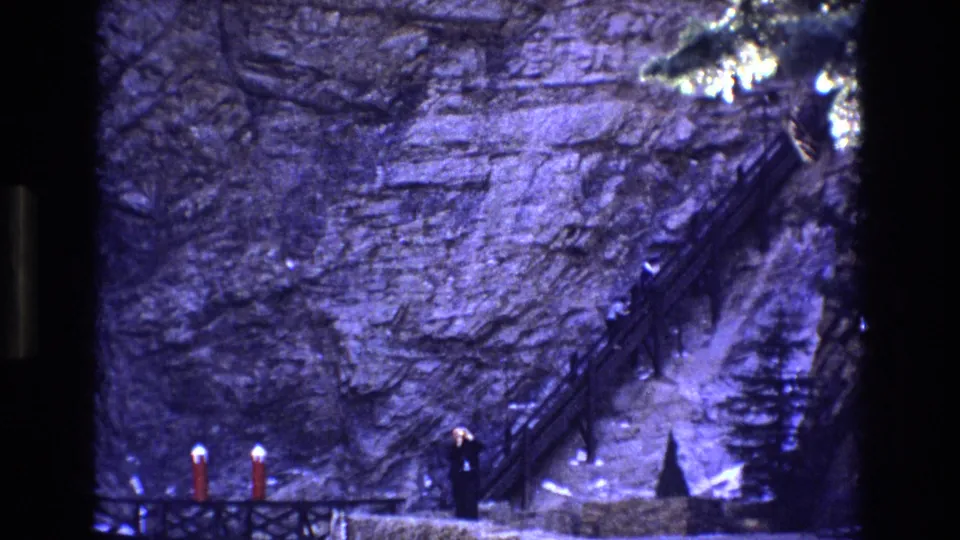 Colorado 1973 home-movie frame with a distant dark-clothed adult standing below a narrow stairway that climbs across a rugged cliff face.