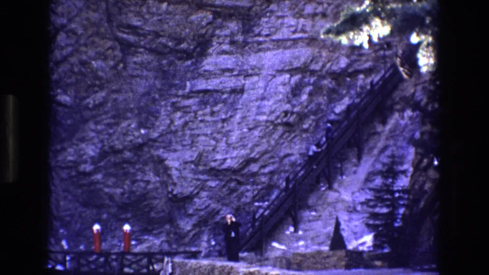 A 1973 Colorado 8mm frame shows a lone adult figure near foreground posts, with a steep staircase cutting upward across the purple-blue cliff wall behind.