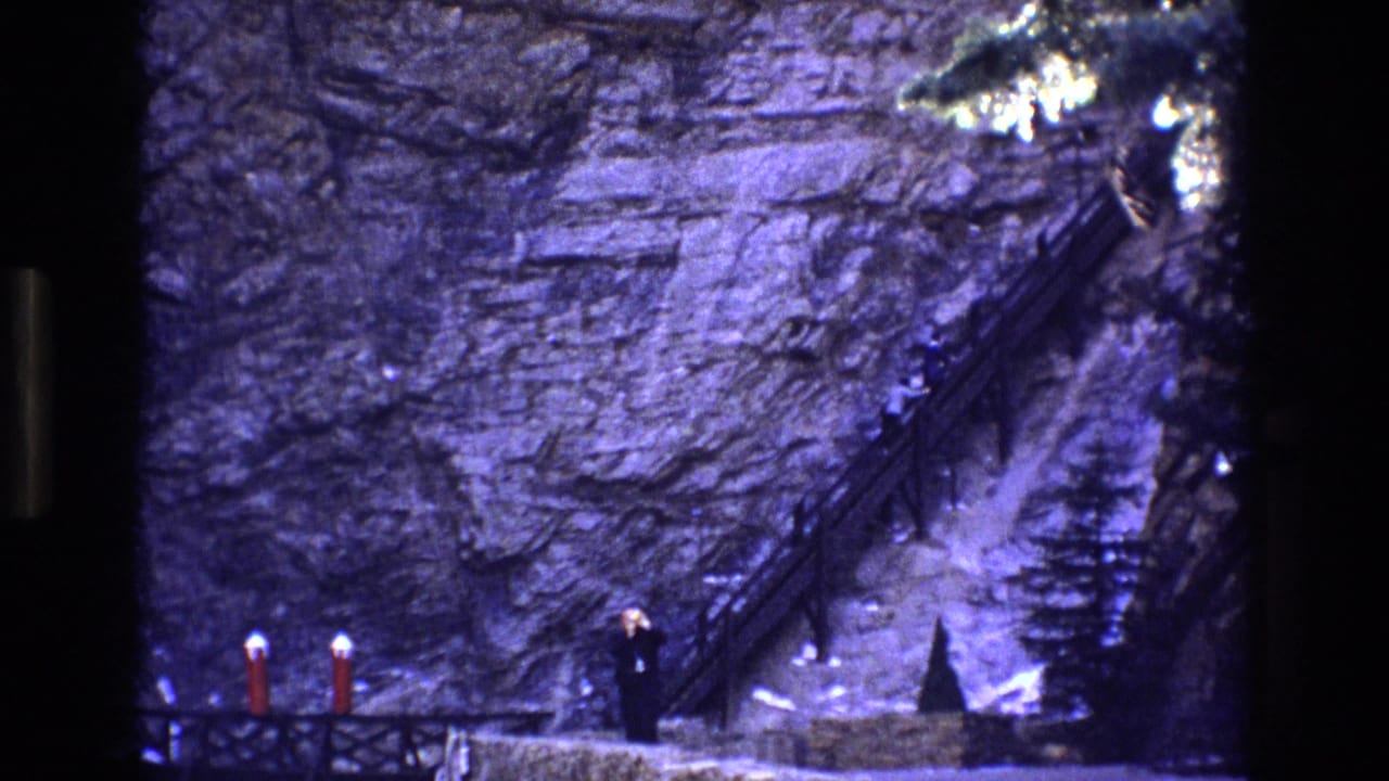 1973 Colorado 8mm frame showing a rocky canyon wall, a narrow stair or path with railings, and a small adult figure near the base of the rocks.