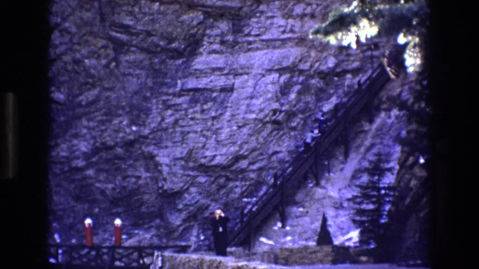 1973 Colorado 8mm frame showing a rocky canyon wall, a narrow stair or path with railings, and a small adult figure near the base of the rocks.