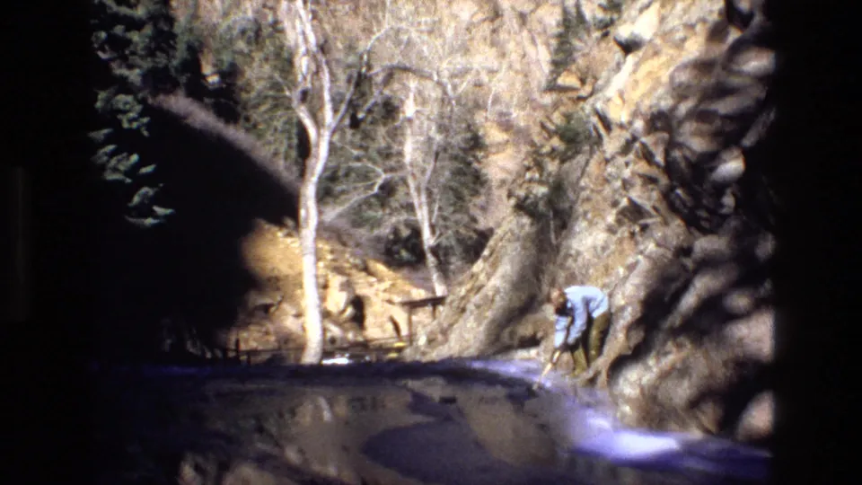 1973 Colorado 8mm frame showing a rocky canyon beside a stream or pool, bare trees, railings, and an adult man standing near the right edge.