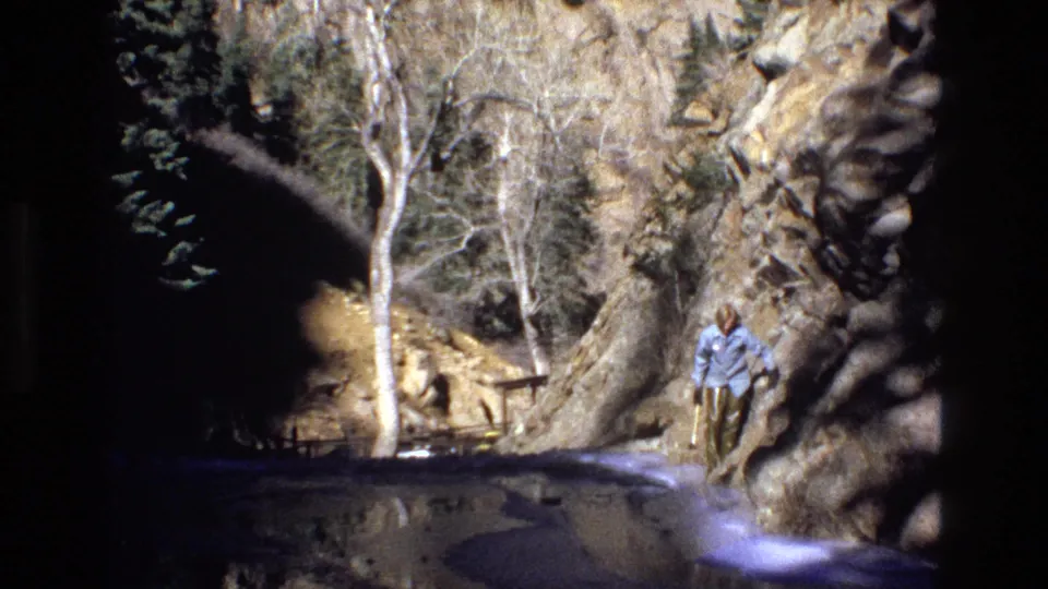 1973 Colorado 8mm frame showing an adult man in a light blue shirt moving along rocks beside canyon water, with bare trees and a small railing behind him.