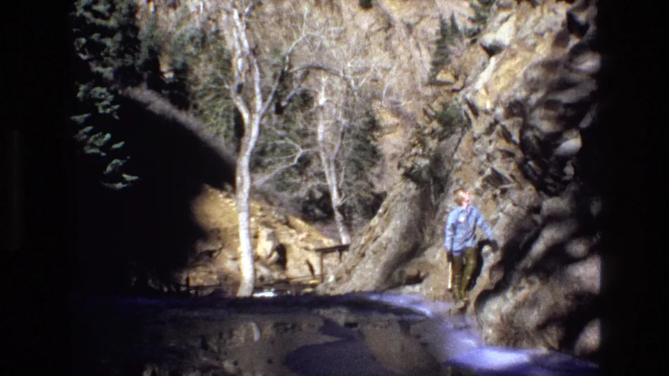 1973 Colorado 8mm frame showing an adult man near a rocky canyon edge with bare trees, a stream or pool, and a small cascade in the foreground.