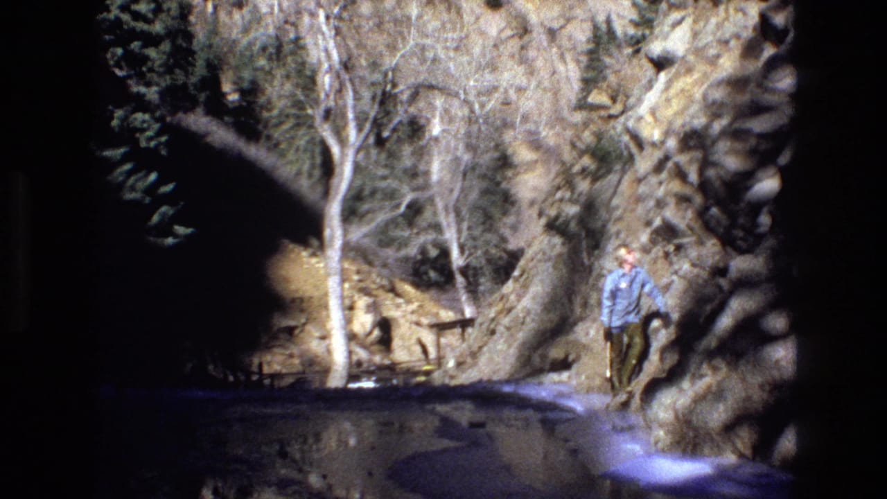 A person in a light blue shirt walks along a shallow mountain stream in a rocky Colorado canyon, seen in 1973 8mm home-movie footage.