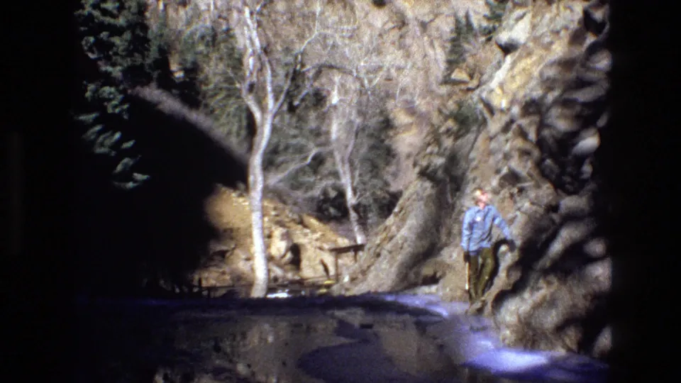 A person in a light blue shirt walks along a shallow mountain stream in a rocky Colorado canyon, seen in 1973 8mm home-movie footage.