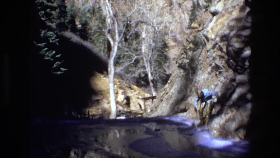 The hiker bends near the stream bank in a rocky Colorado canyon, with bare trees and strong sunlight visible in the 8mm frame.
