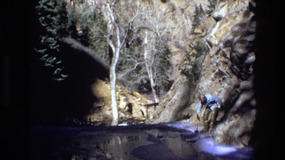 A person in a light blue shirt bends beside shallow water in a rugged Colorado canyon, captured as grainy 1973 8mm home-movie footage.