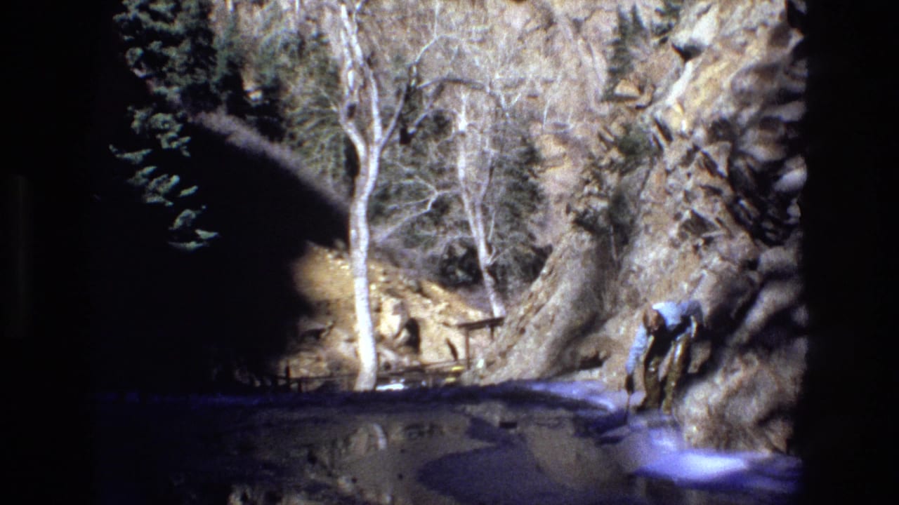1973 Colorado 8mm frame showing a man in a light shirt bending near a rocky stream beneath bare trees and steep rock walls.