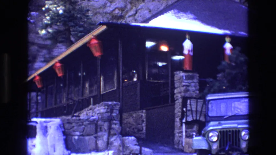 1973 Colorado 8mm frame looking along a snow-covered mountain cabin porch with red lanterns, stone base, dark siding, and part of a Jeep-style vehicle at the edge.