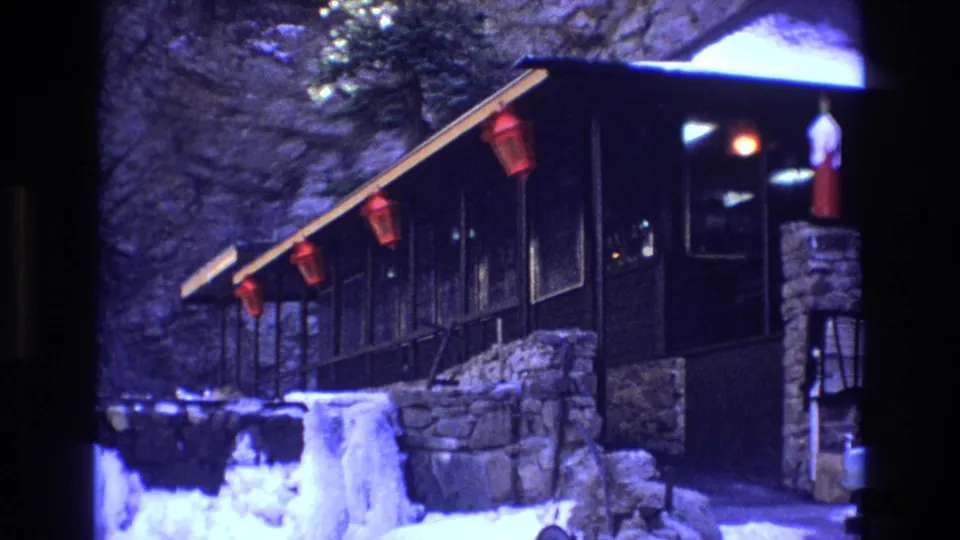 1973 Colorado home-movie frame showing the snowy exterior of a dark wood mountain cabin with red lanterns, stonework, warm interior light, and a rocky cliff backdrop.