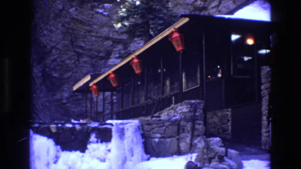 1973 Colorado 8mm frame showing a dark wooden mountain lodge porch with red lanterns, stonework, snowbanks, and ice in the foreground.