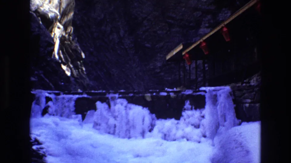 1973 Colorado 8mm frame showing a wider snowy foreground, icy formations, a rocky cliff, and the lodge porch with red lanterns receding at the right edge.