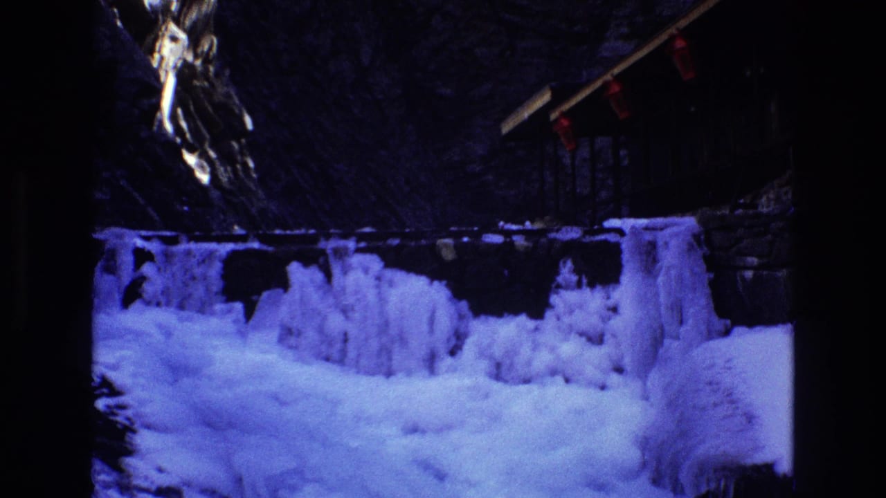 1973 Colorado 8mm frame showing a blue-toned frozen waterfall, snowy bank, dark rocky wall, and small red lights near a wooden structure above the ice.