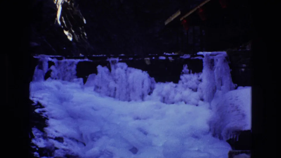 1973 Colorado 8mm frame showing a frozen cascade with heavy snow and ice in the foreground, dark rock behind, and red lights partly visible overhead.