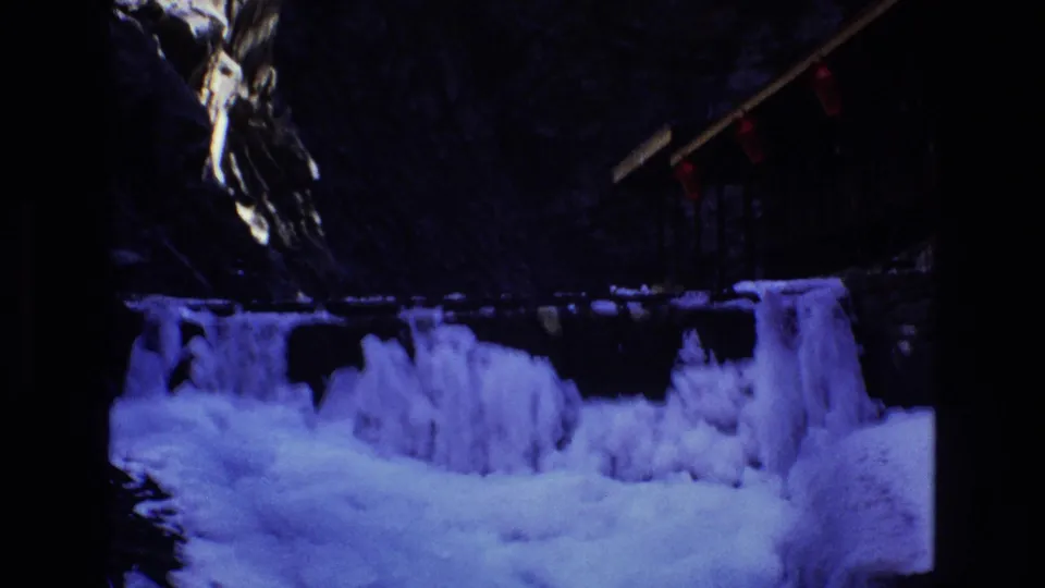 1973 Colorado 8mm frame capturing the frozen waterfall from a slightly shifted view, with blue ice, dark cliffs, red lights, and a small wooden structure overhead.