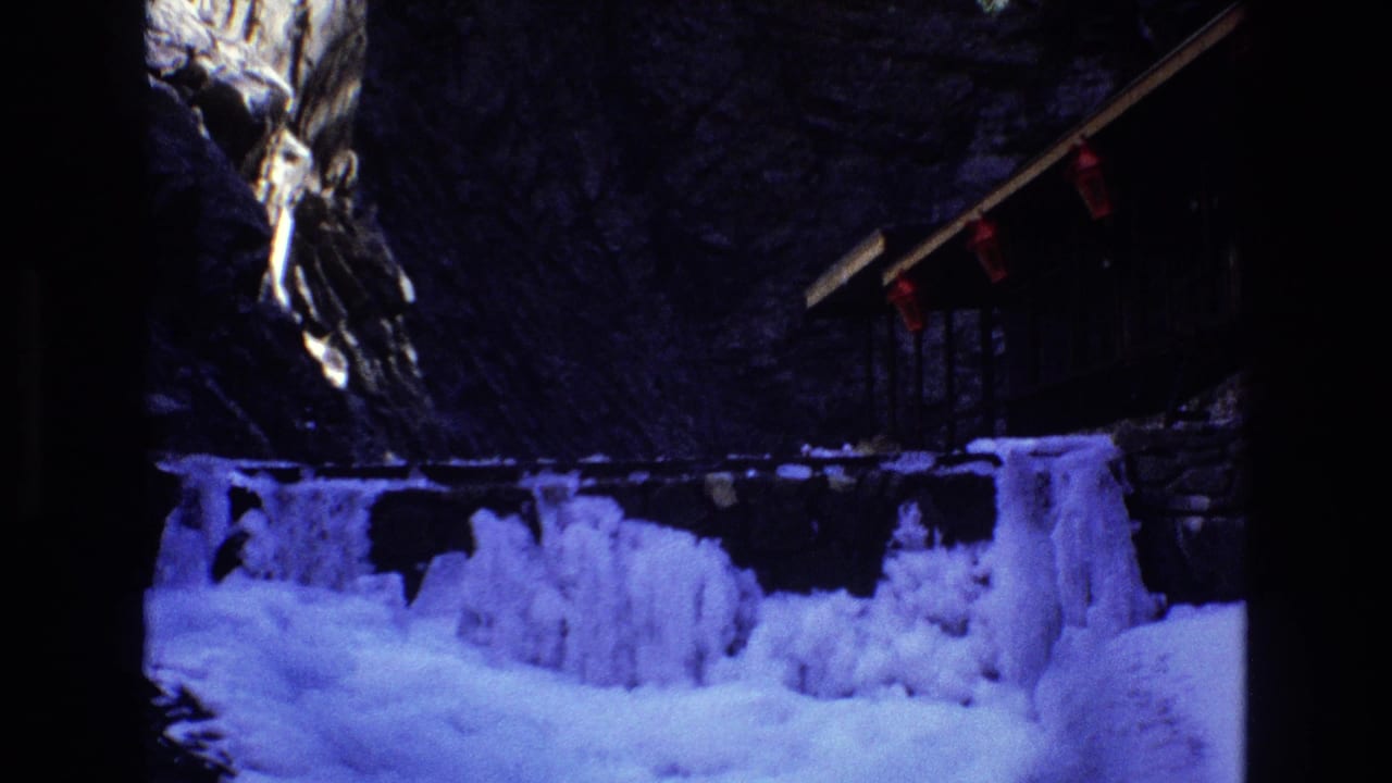 1973 Colorado 8mm frame showing snow and ice along a mountain stream beneath a rocky cliffside wooden structure with red hanging fixtures.