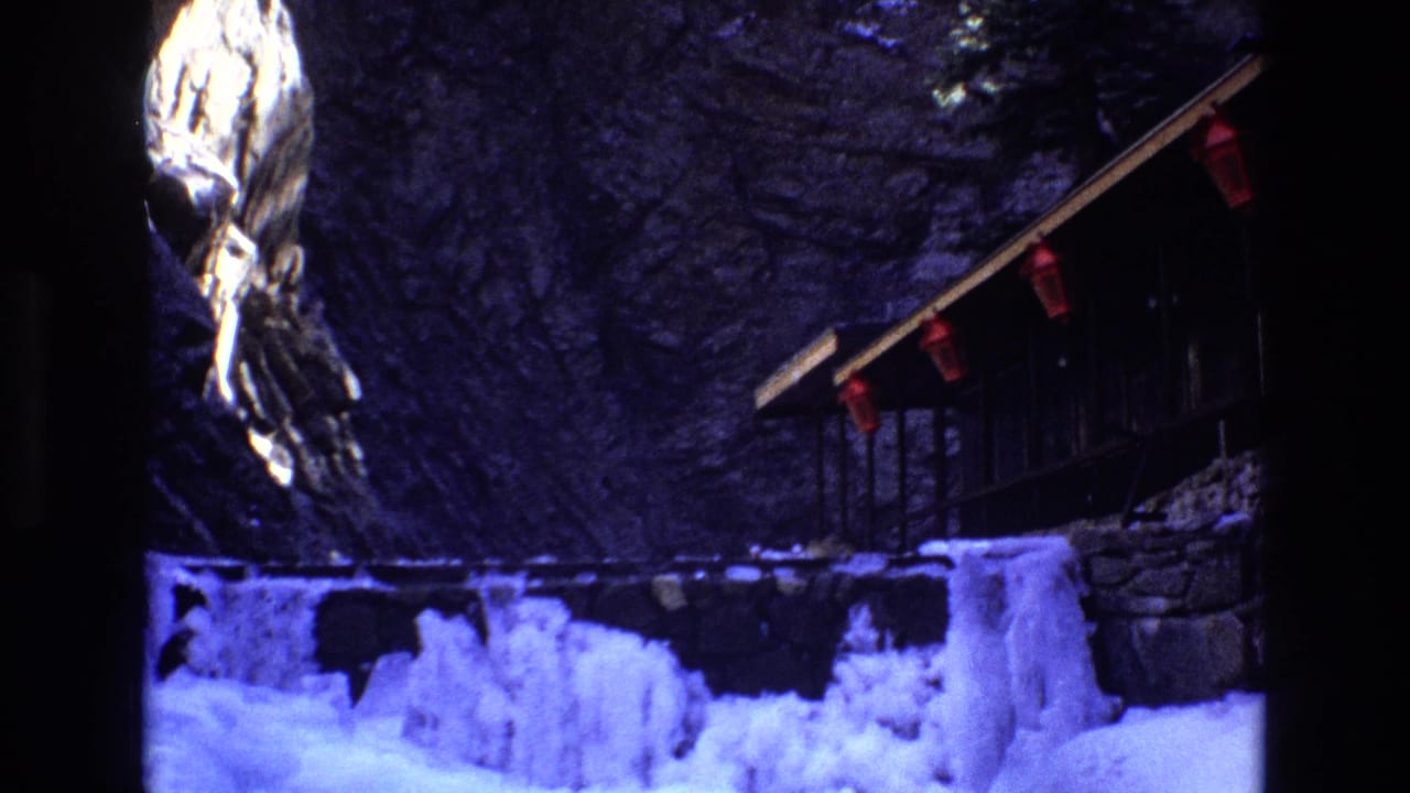 Snowy frozen stream and cliffside wooden structure seen through dark film-frame edges.