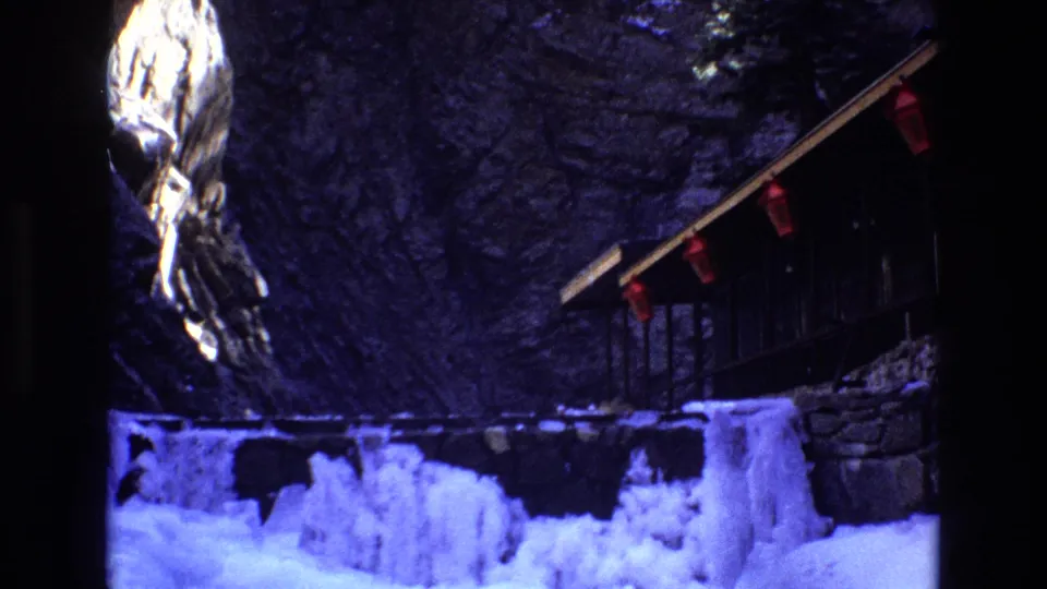 1973 Colorado home-movie frame with blue-toned ice, rocky canyon wall, and a wooden cliffside structure with small red fixtures.