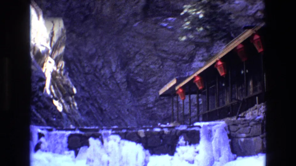1973 Colorado 8mm frame showing a cliffside wooden structure with red hanging fixtures above an icy stream and snow-covered rocks.