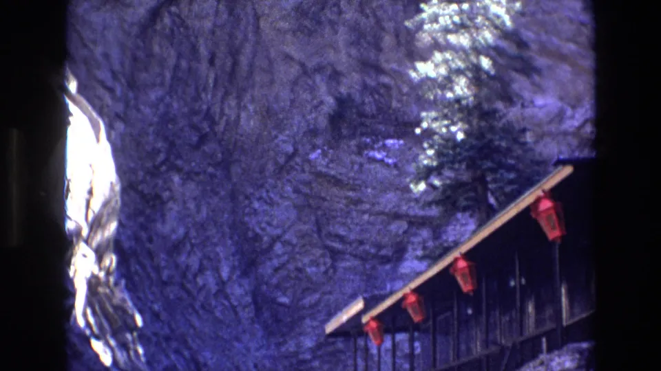 1973 Colorado frame with a dark rock face, snow and ice patches, and part of a wooden cliffside structure carrying red hanging fixtures.