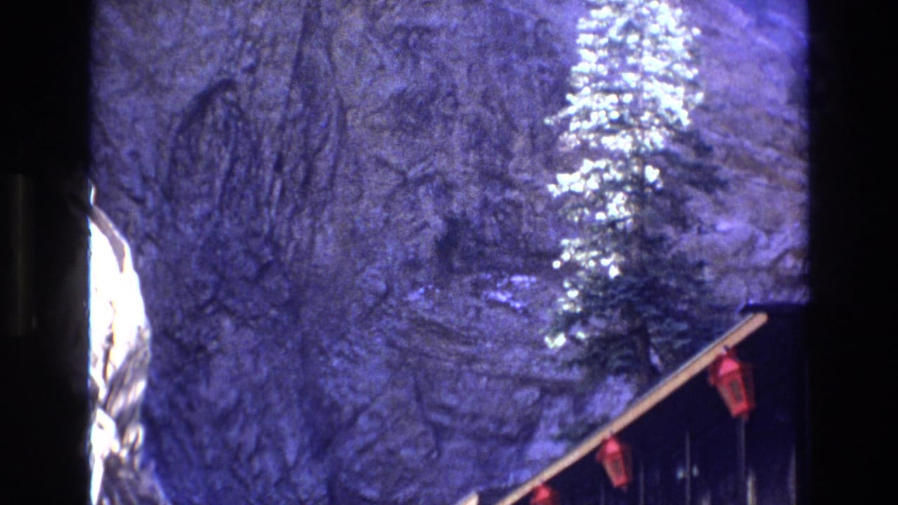 1973 Colorado 8mm frame showing a rocky cliff face, a tall conifer, and the edge of a dark wooden scenic overlook railing with red details.