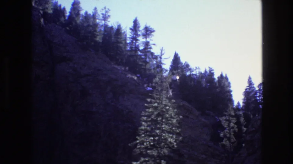 1973 Colorado 8mm frame of a shadowed rocky mountainside with conifers rising toward a bright sky and forested upper ridge.