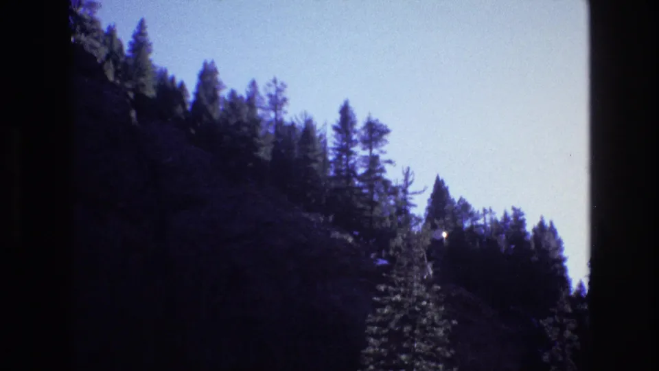 1973 Colorado 8mm frame of a rocky, pine-lined valley wall with a small bright point visible through the trees.