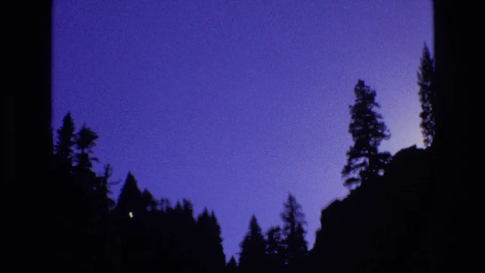 1973 Colorado 8mm frame showing dark pine silhouettes rising against a blue-purple sky in a shaded mountain valley.
