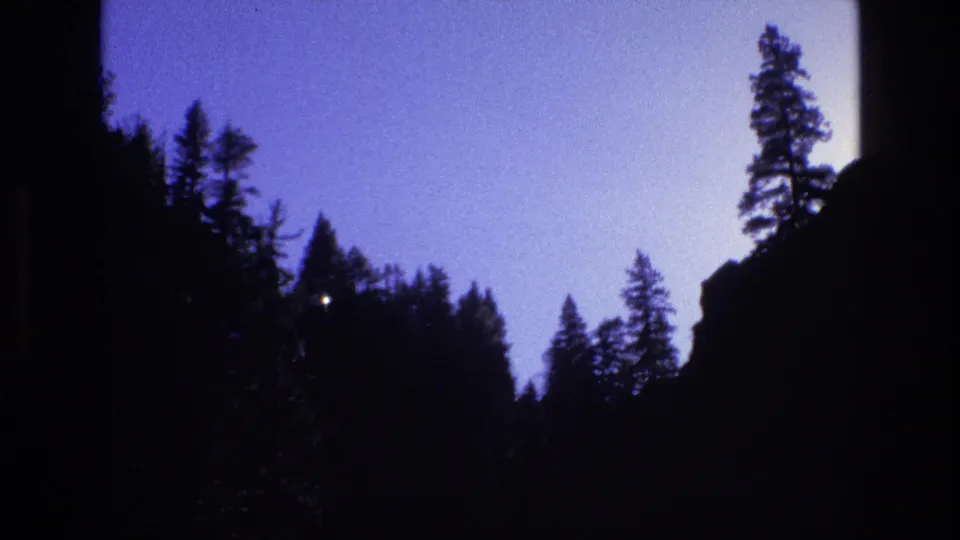 1973 Colorado 8mm frame of a shadowed pine-filled mountain valley between dark rocky edges beneath a pale evening or morning sky.
