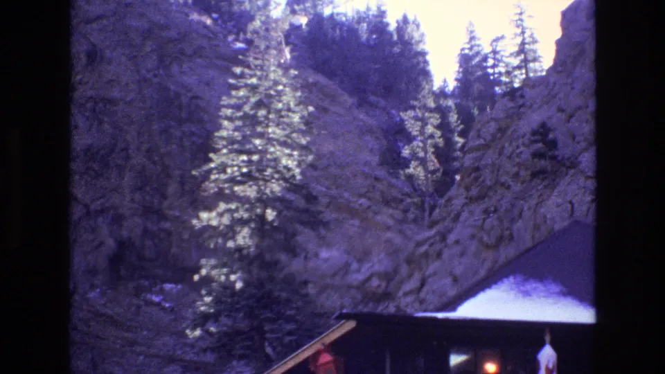 1973 Colorado 8mm frame showing a pine-lined rock wall above the edge of a hut-like sloped roof in a canyon setting.
