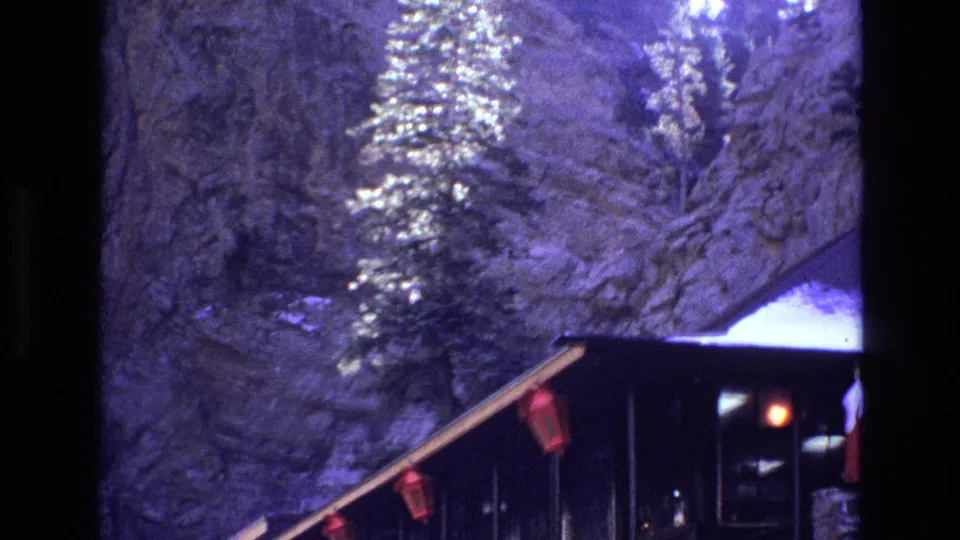 1973 Colorado 8mm frame showing a rustic hut-like structure with red lanterns and a small warm light beneath steep canyon rock and conifer trees.